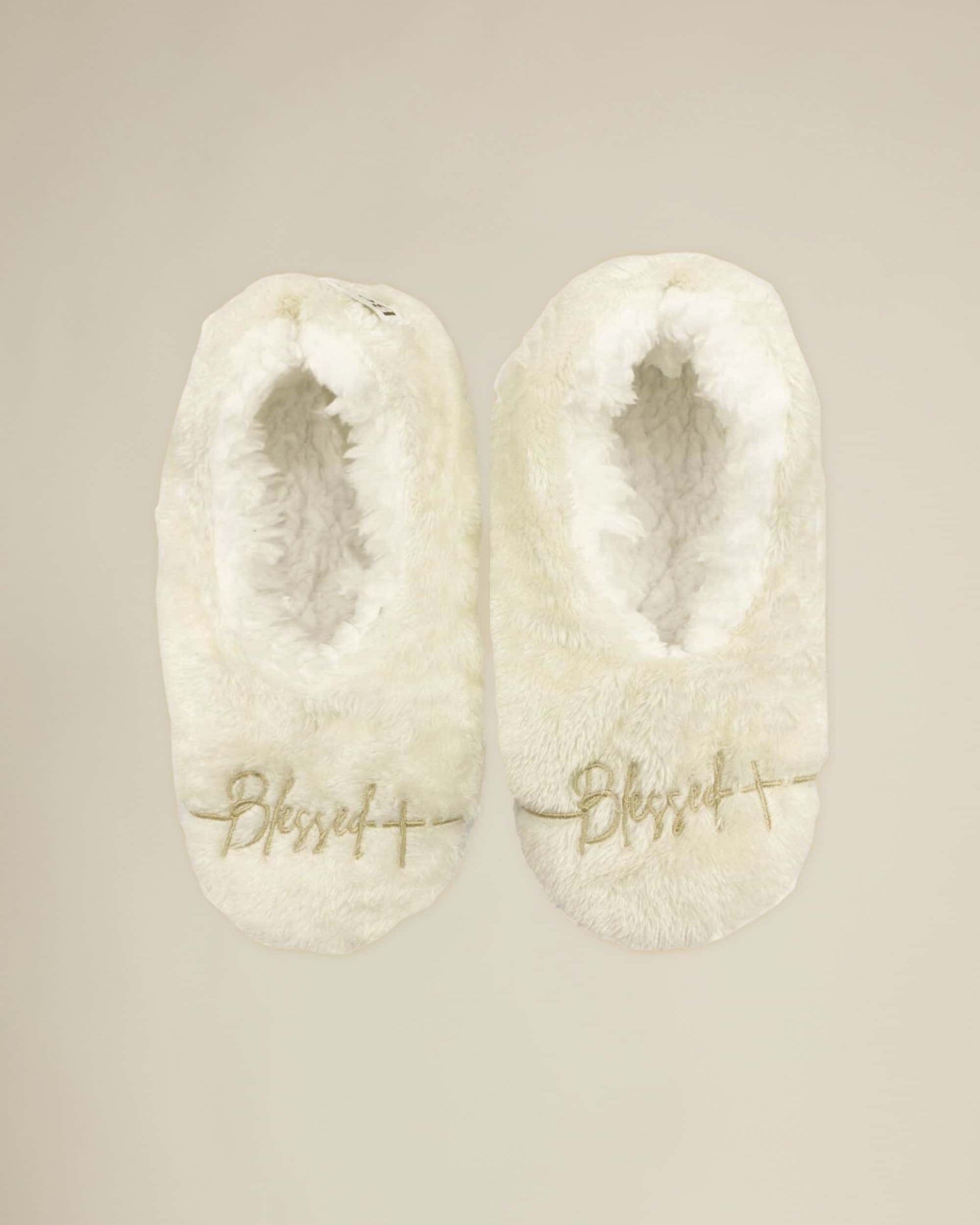 A pair of white plush slippers with 'Blessed' written on the top in a golden script.