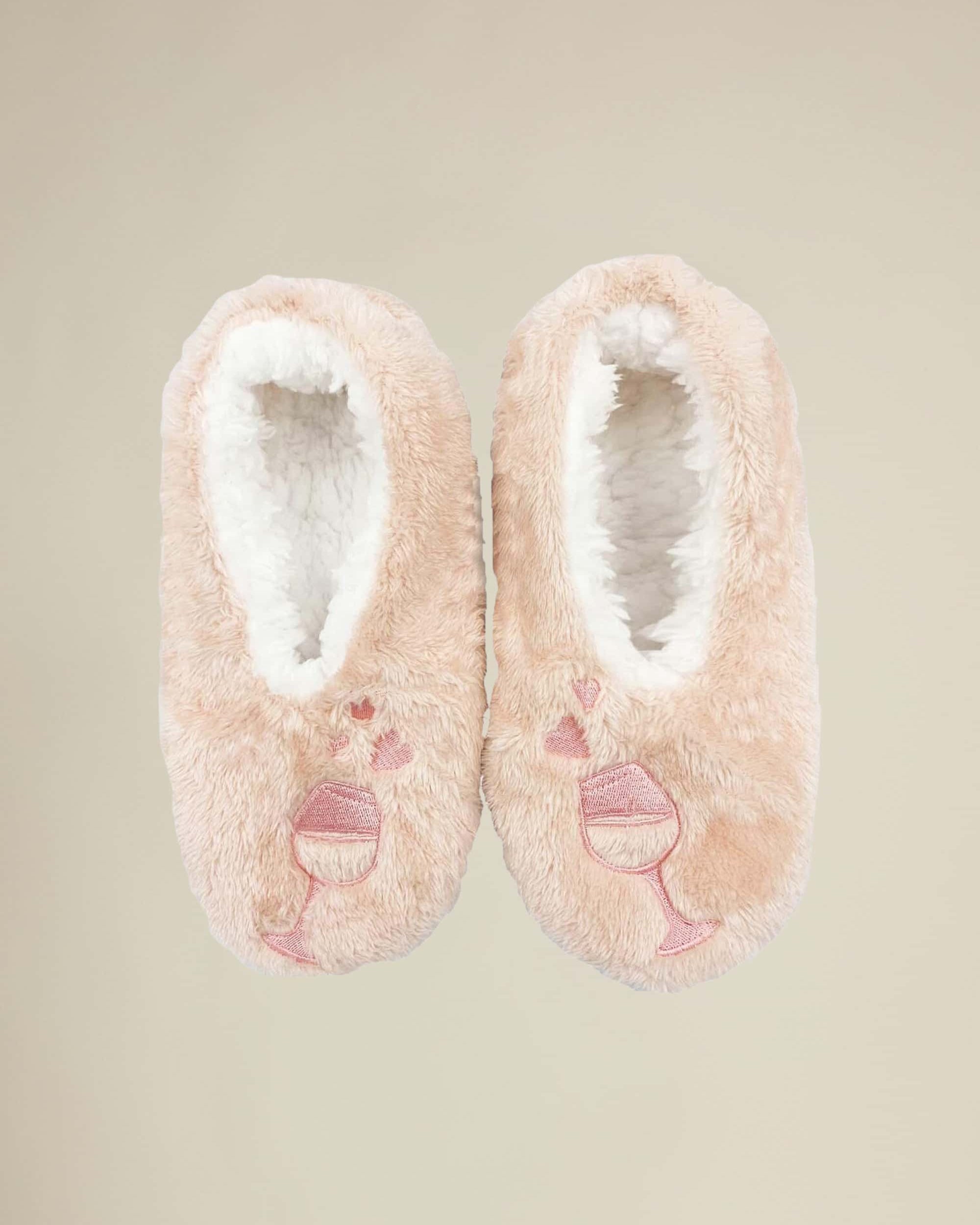 A pair of pink plush slippers designed to look like wine glasses with hearts, with the phrase 'Wine a little. Laugh a lot.