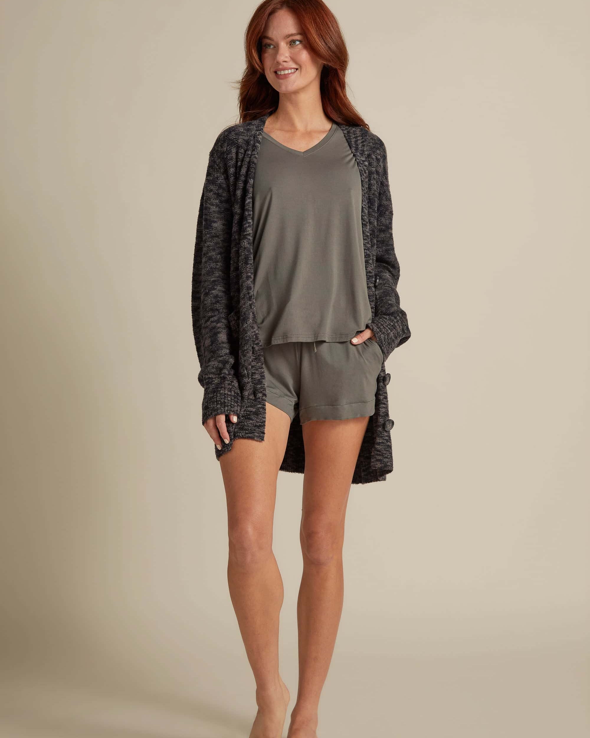 A female model wearing a gray button-up cardigan with side pockets over a matching V-neck shirt, paired with shorts.