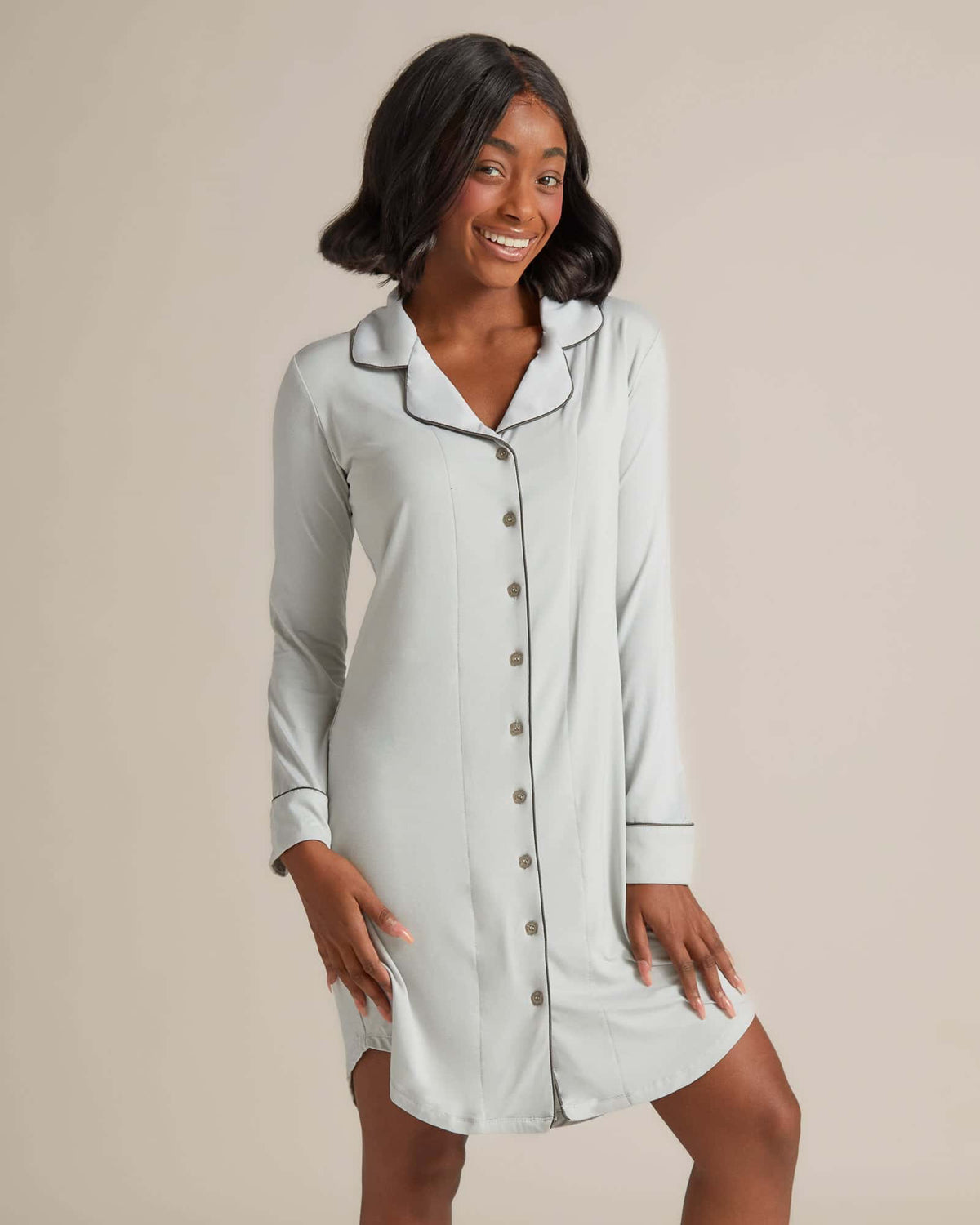 A woman standing and wearing a grey nightshirt with long sleeves and button-up front.