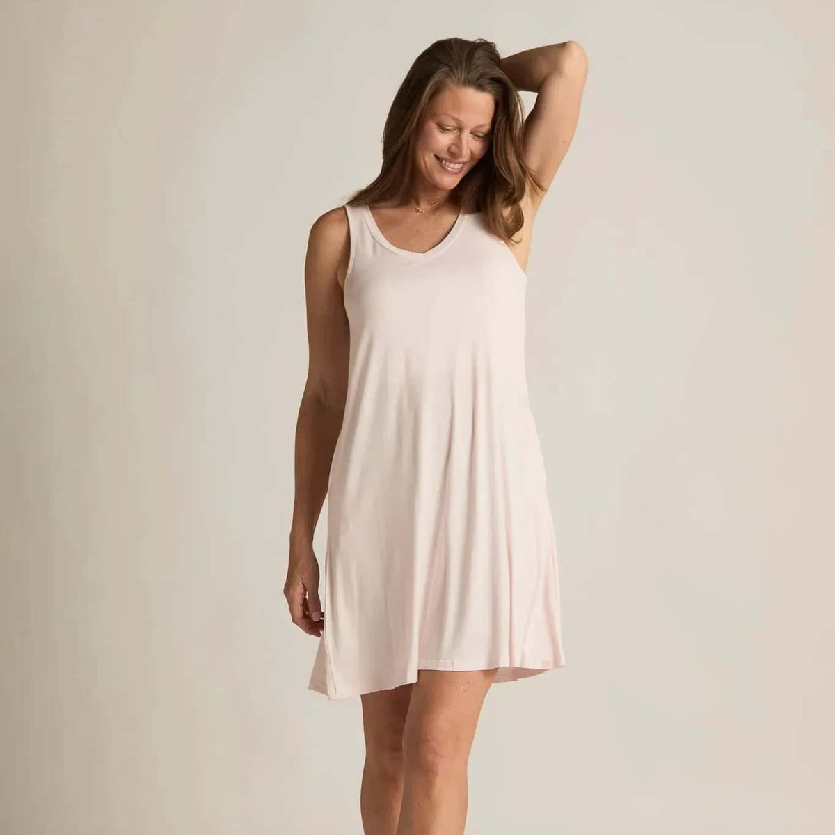 A woman standing, wearing a sleeveless pink nightshirt with a V-neckline in front and back, side pockets, and an A-line hem.