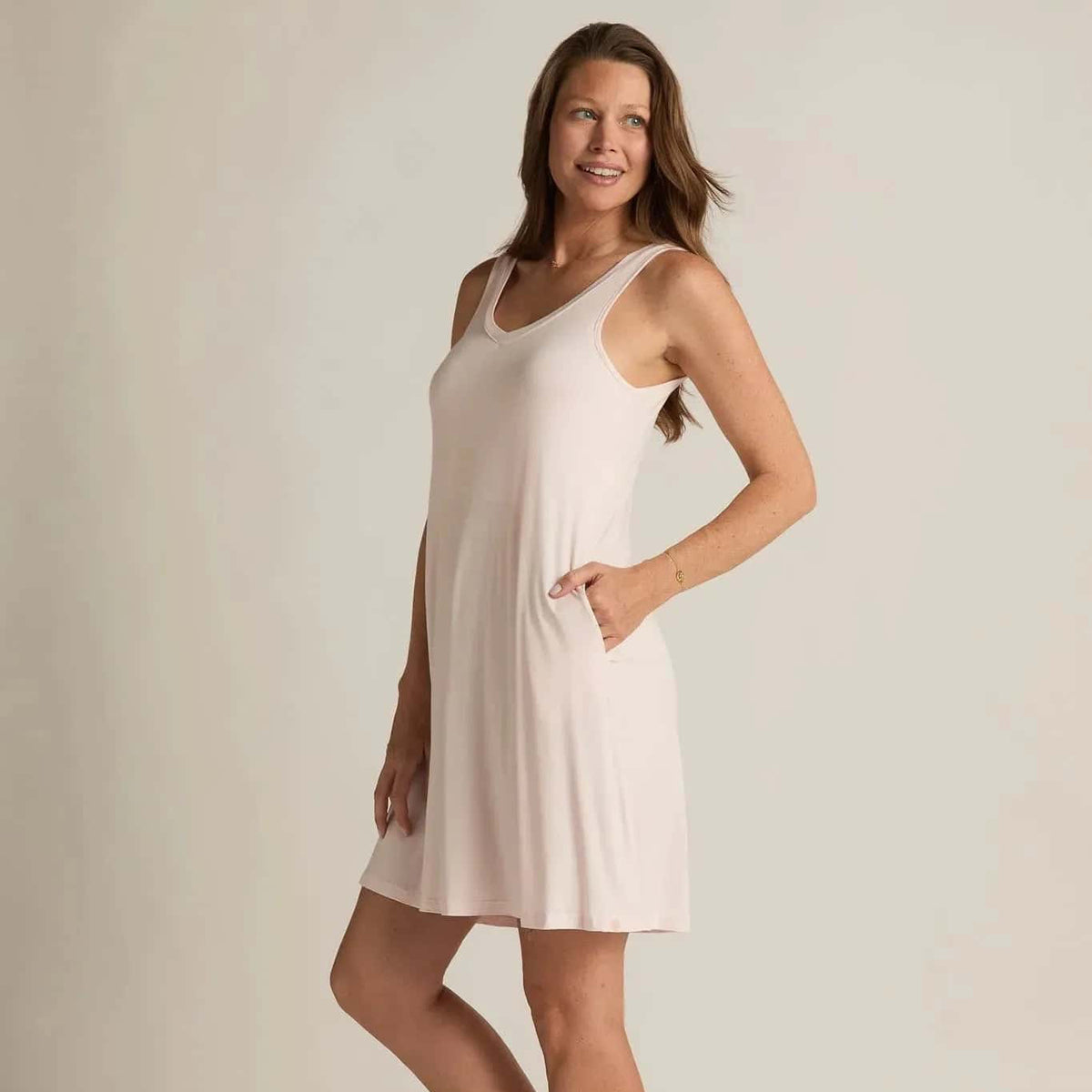 A woman standing, wearing a sleeveless pink nightshirt with a V-neckline in front and back, side pockets, and an A-line hem.