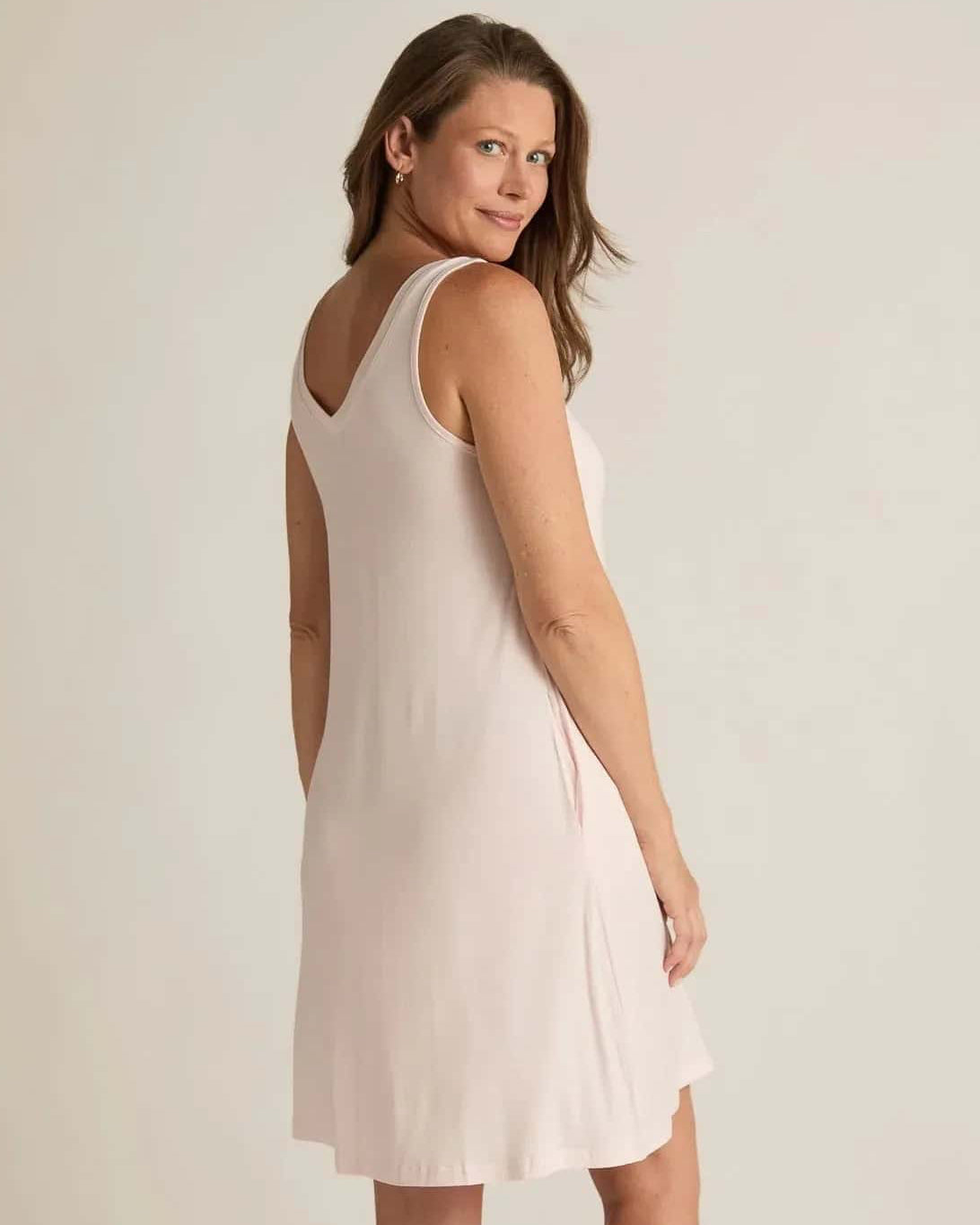 A woman standing, wearing a sleeveless pink nightshirt with a V-neckline in front and back, side pockets, and an A-line hem.