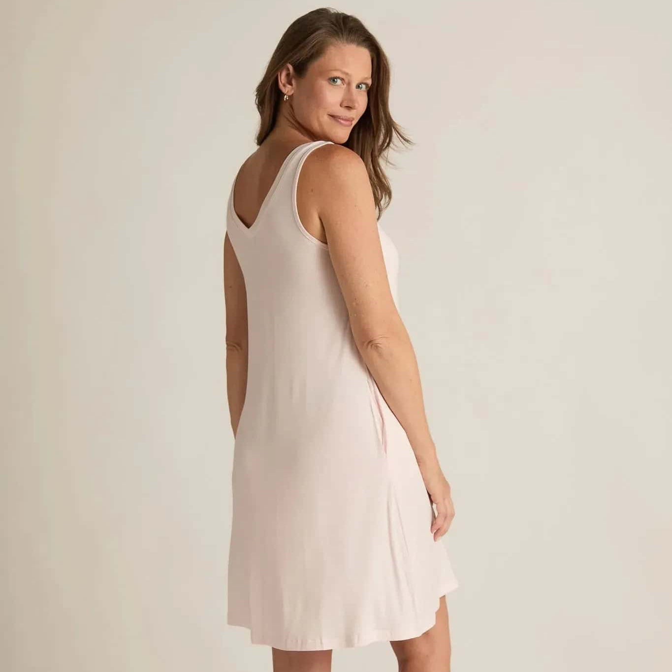A woman standing, wearing a sleeveless pink nightshirt with a V-neckline in front and back, side pockets, and an A-line hem.