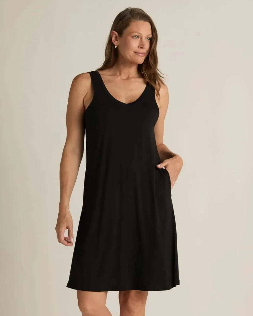 A woman standing, wearing a sleeveless black nightshirt with a V-neckline in front and back, side pockets, and an A-line hem.