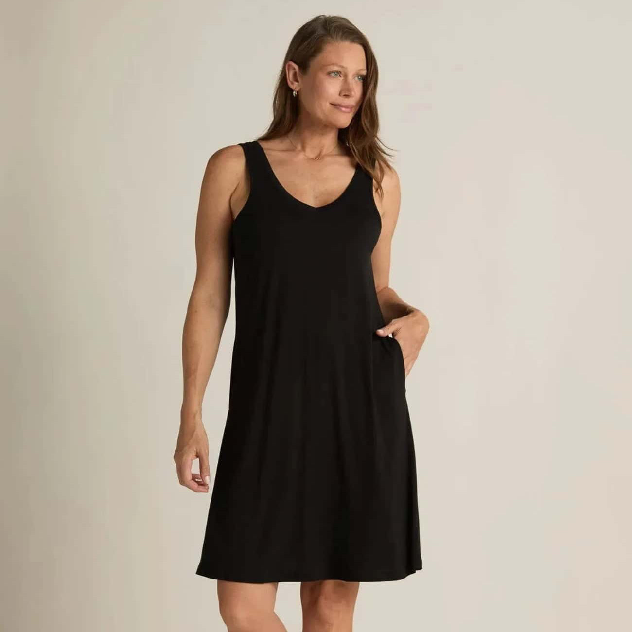 A woman standing, wearing a sleeveless black nightshirt with a V-neckline in front and back, side pockets, and an A-line hem.