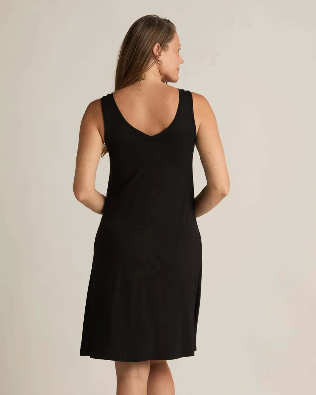 A woman standing, wearing a sleeveless black nightshirt with a V-neckline in front and back, side pockets, and an A-line hem.