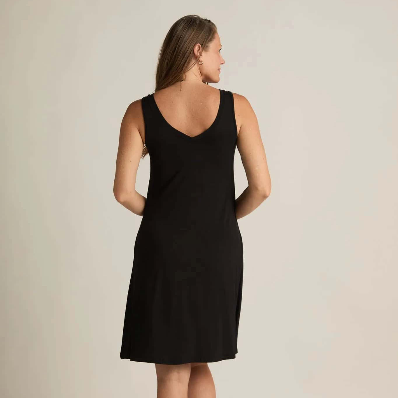 A woman standing, wearing a sleeveless black nightshirt with a V-neckline in front and back, side pockets, and an A-line hem.