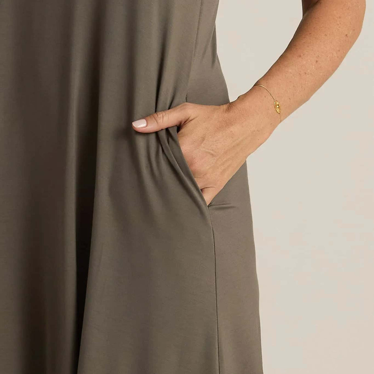 A woman standing, wearing a sleeveless grey nightshirt with a V-neckline in front and back, side pockets, and an A-line hem.