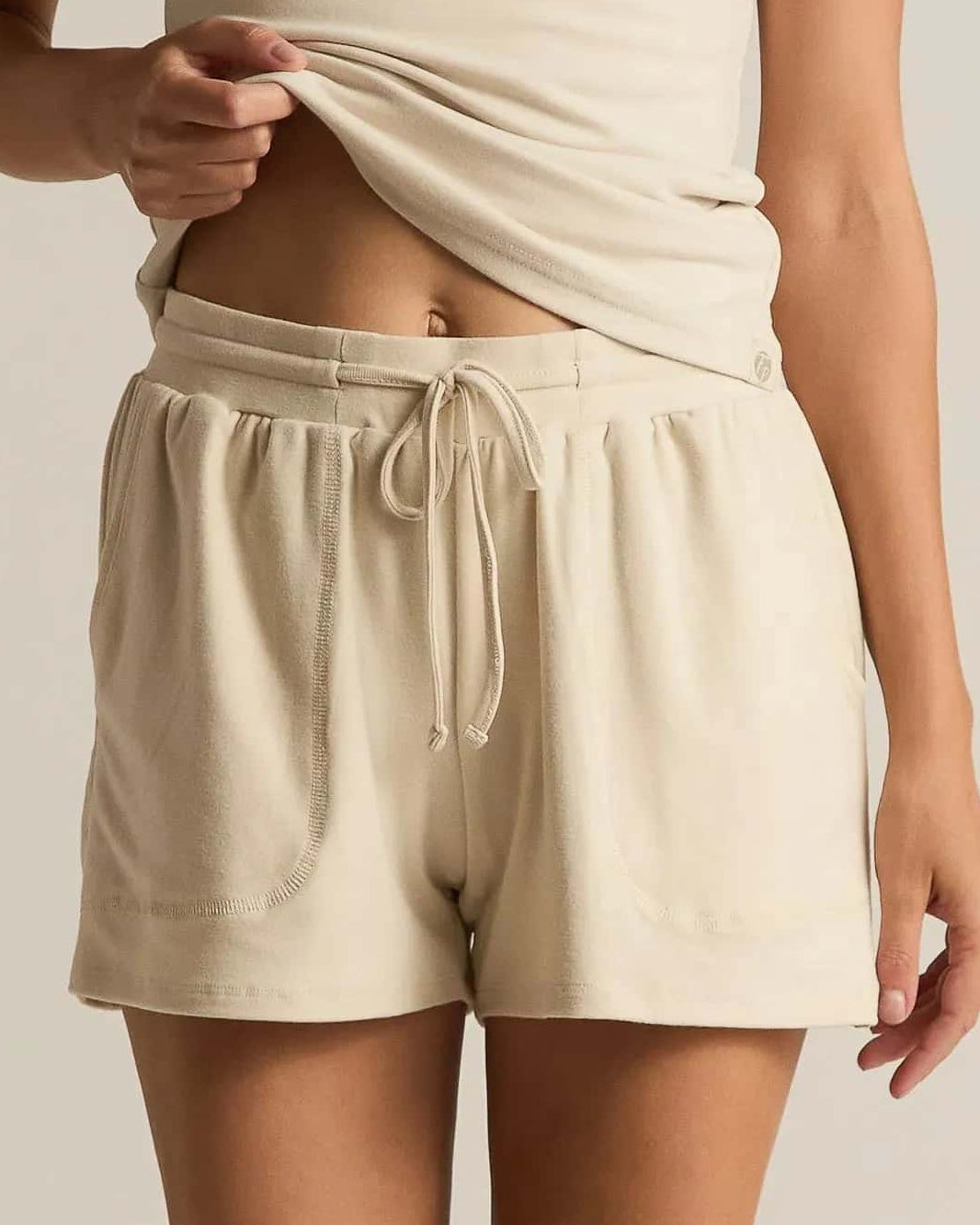 A woman wearing beige short lounge shorts with a drawstring waistband and a matching tank top