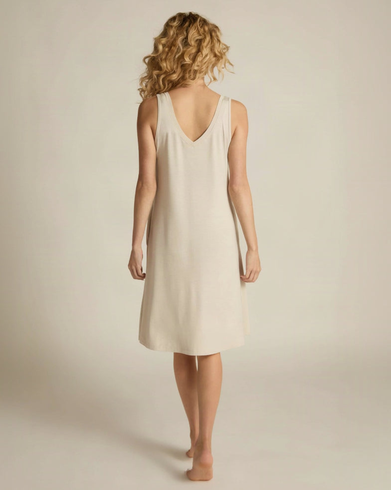 A woman standing, wearing a sleeveless beige nightshirt with a V-neckline in front and back, side pockets, and an A-line hem.