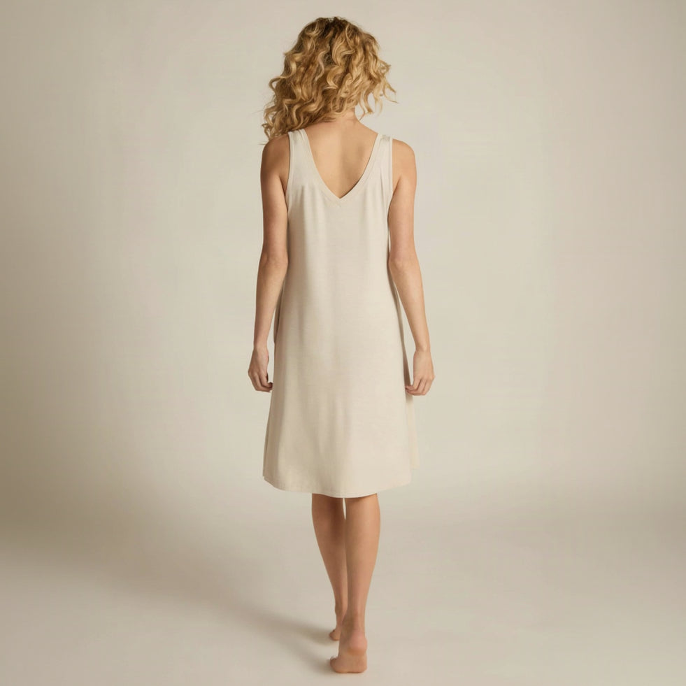 A woman standing, wearing a sleeveless beige nightshirt with a V-neckline in front and back, side pockets, and an A-line hem.