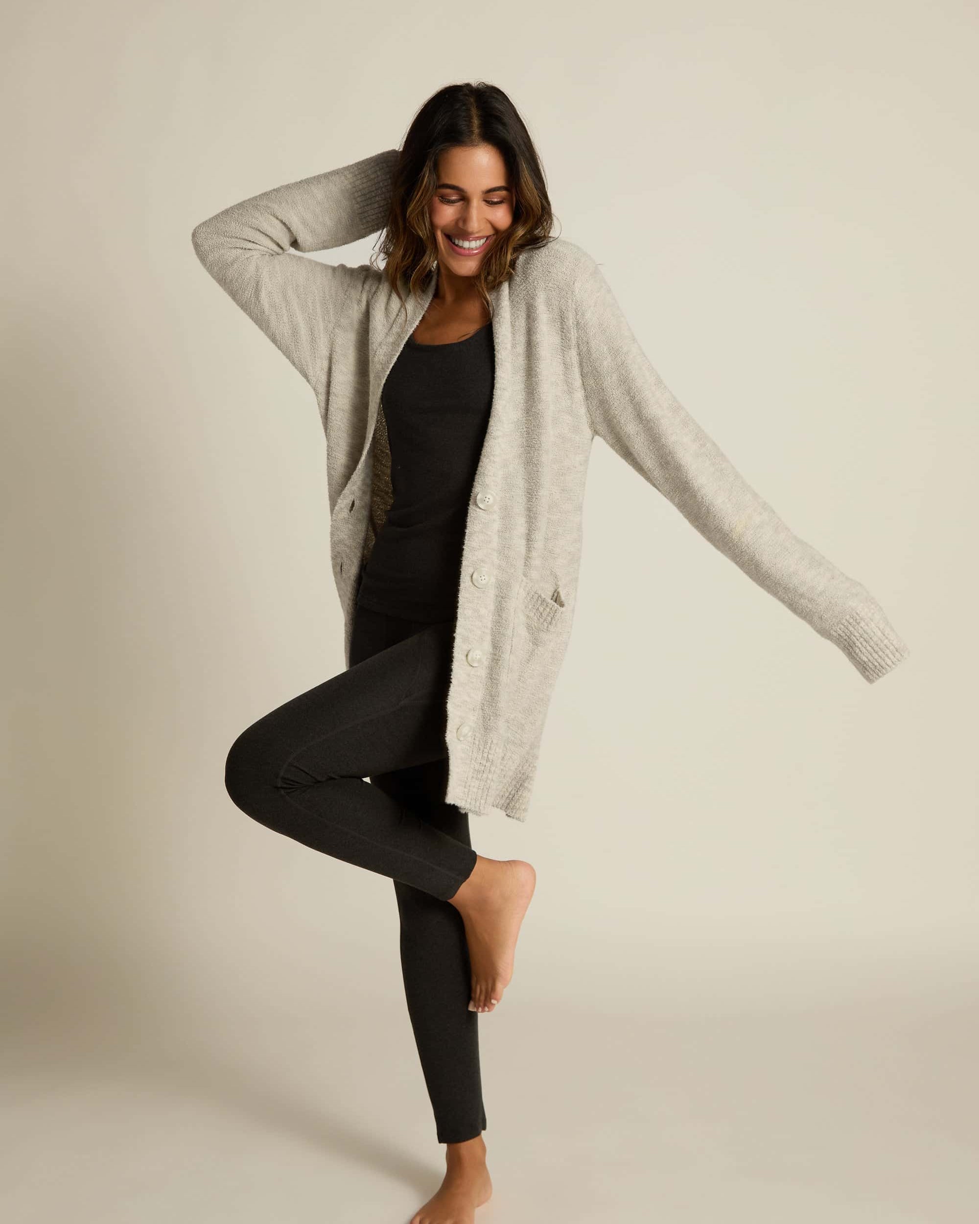 A female model wearing a gray button-up cardigan with side pockets over a matching black shirt and leggings. She stands on one leg with arms posed