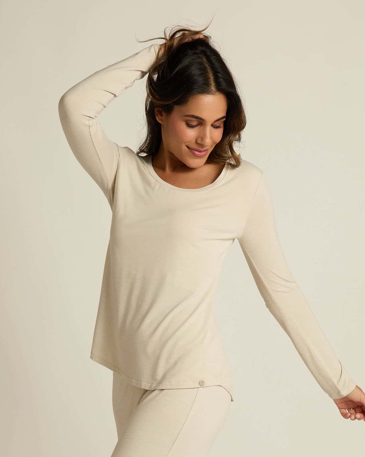 A woman wearing a beige long sleeve top and matching pants, standing against a plain background