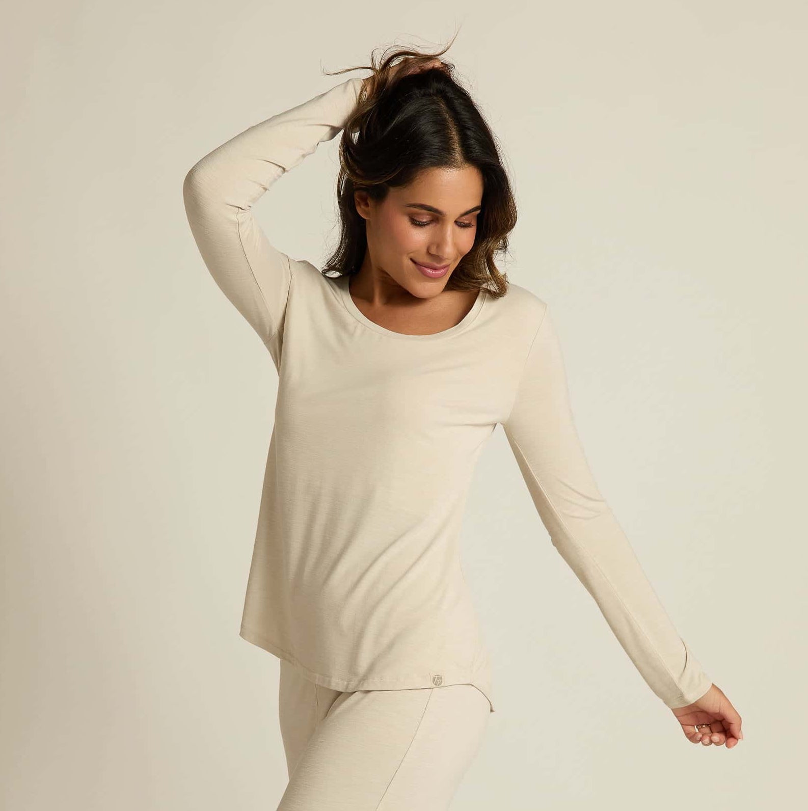 A woman wearing a beige long sleeve top and matching pants, standing against a plain background