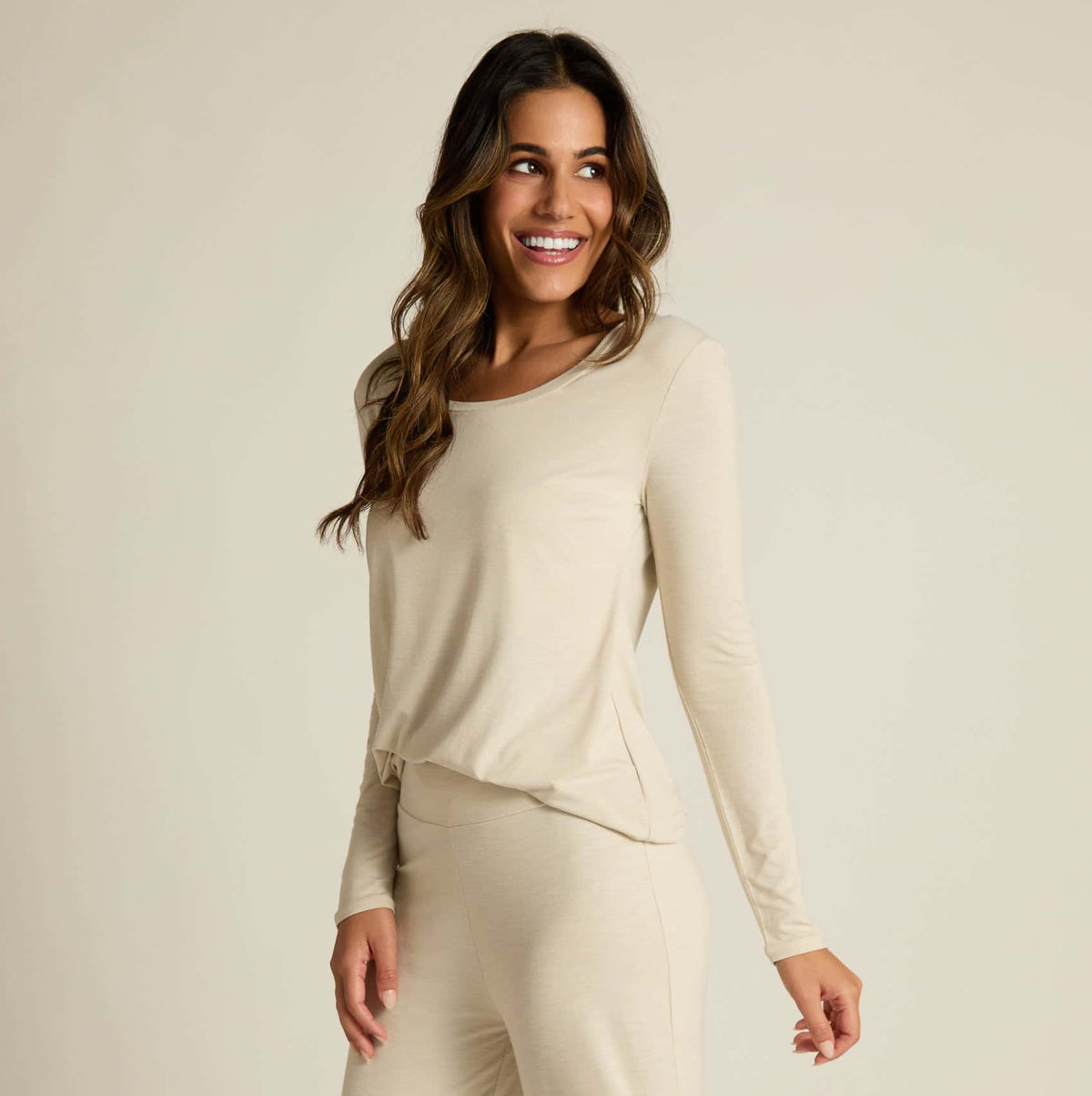A woman wearing a beige long sleeve top and matching pants, standing against a plain background