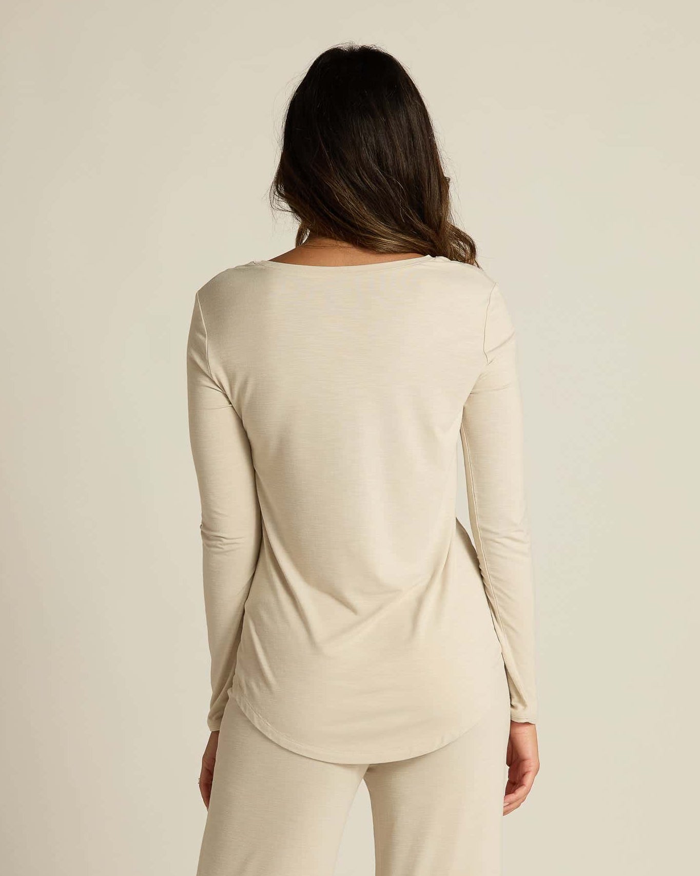 A woman wearing a beige long sleeve top and matching pants, standing against a plain background