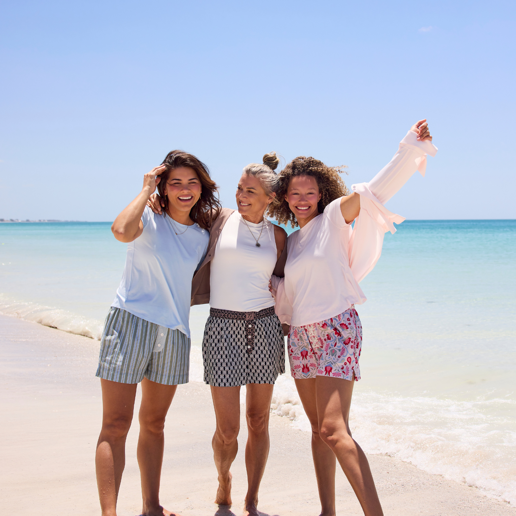 Three women on a sunny beach with the ocean and blue sky in the background, wearing Faceplant Dreams' new cotton print shorts and the softest breathable loungewear tops in white, sky blue, and pink.