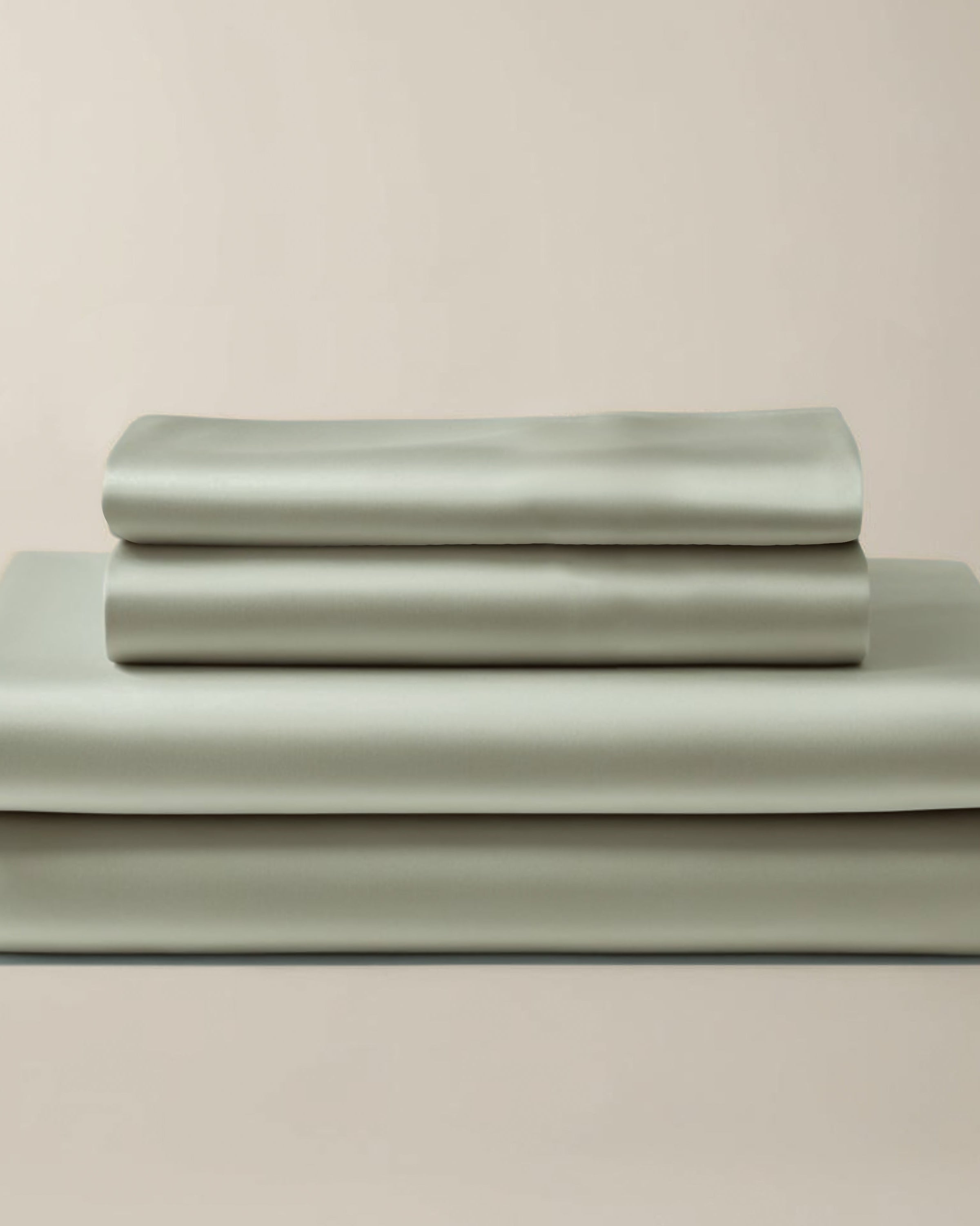Set of satin sage green bedding sheets stacked on a beige background