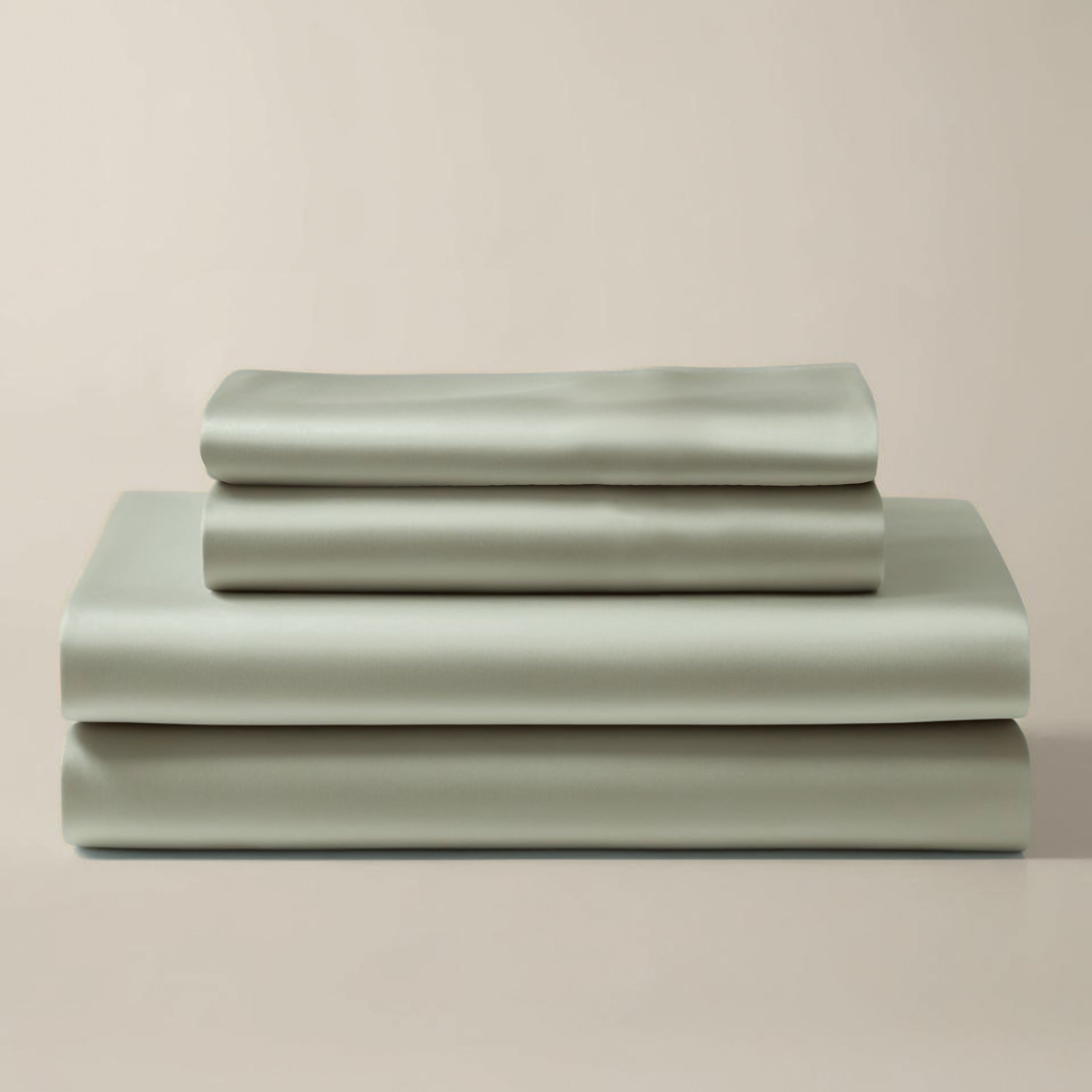 Set of satin sage green bedding sheets stacked on a beige background