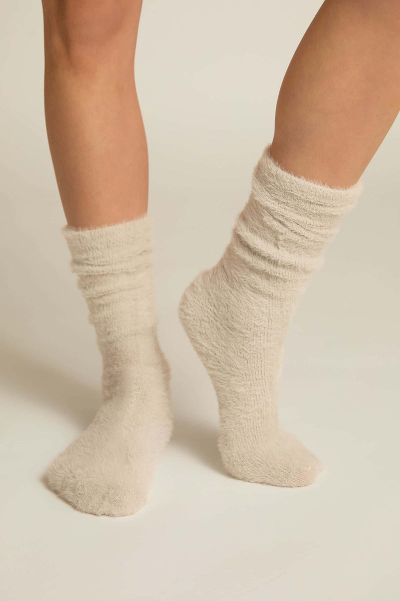 A pair of beige fuzzy lounge socks worn on human legs, standing against a white background.