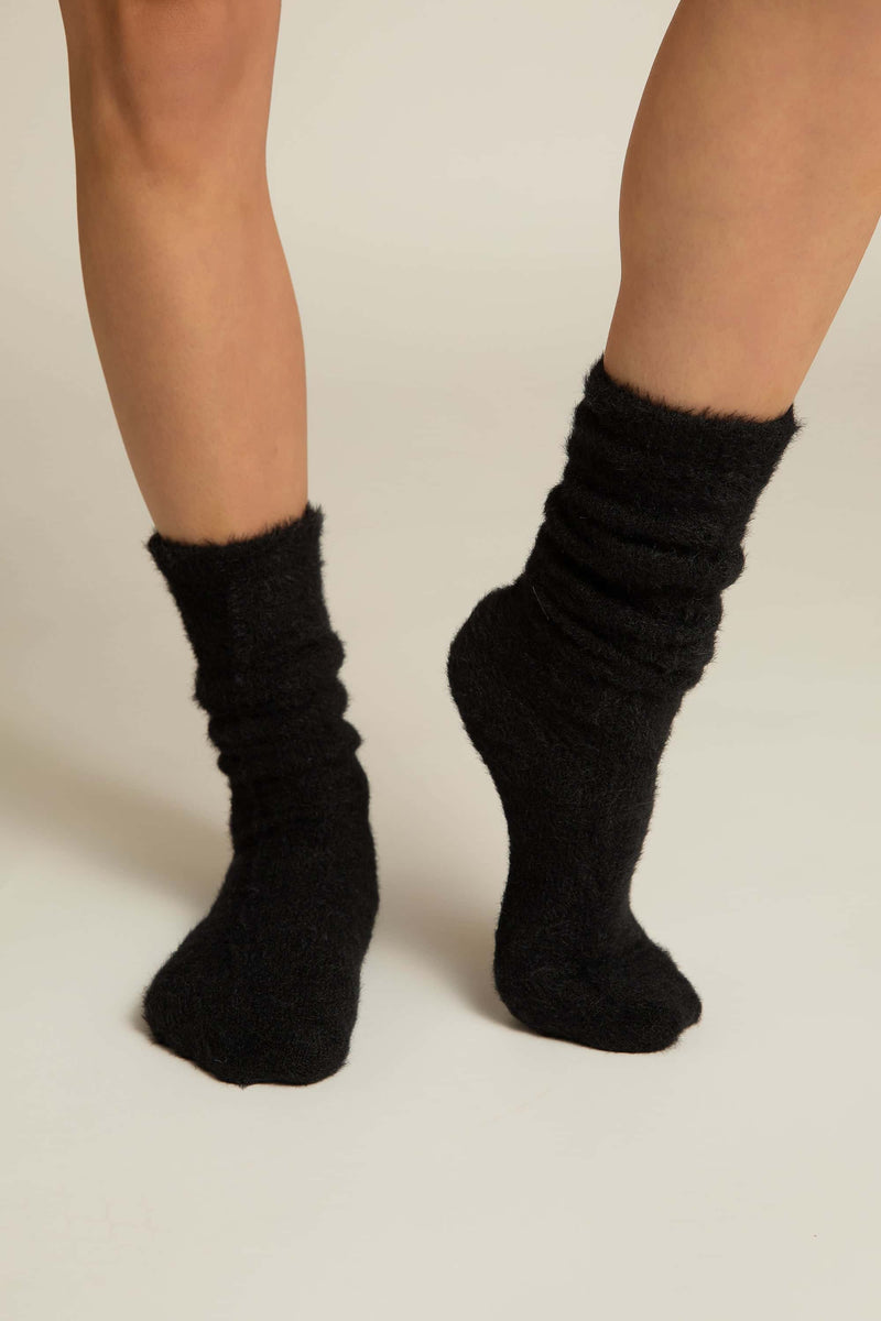 A pair of black fuzzy lounge socks worn on human legs, standing against a white background.