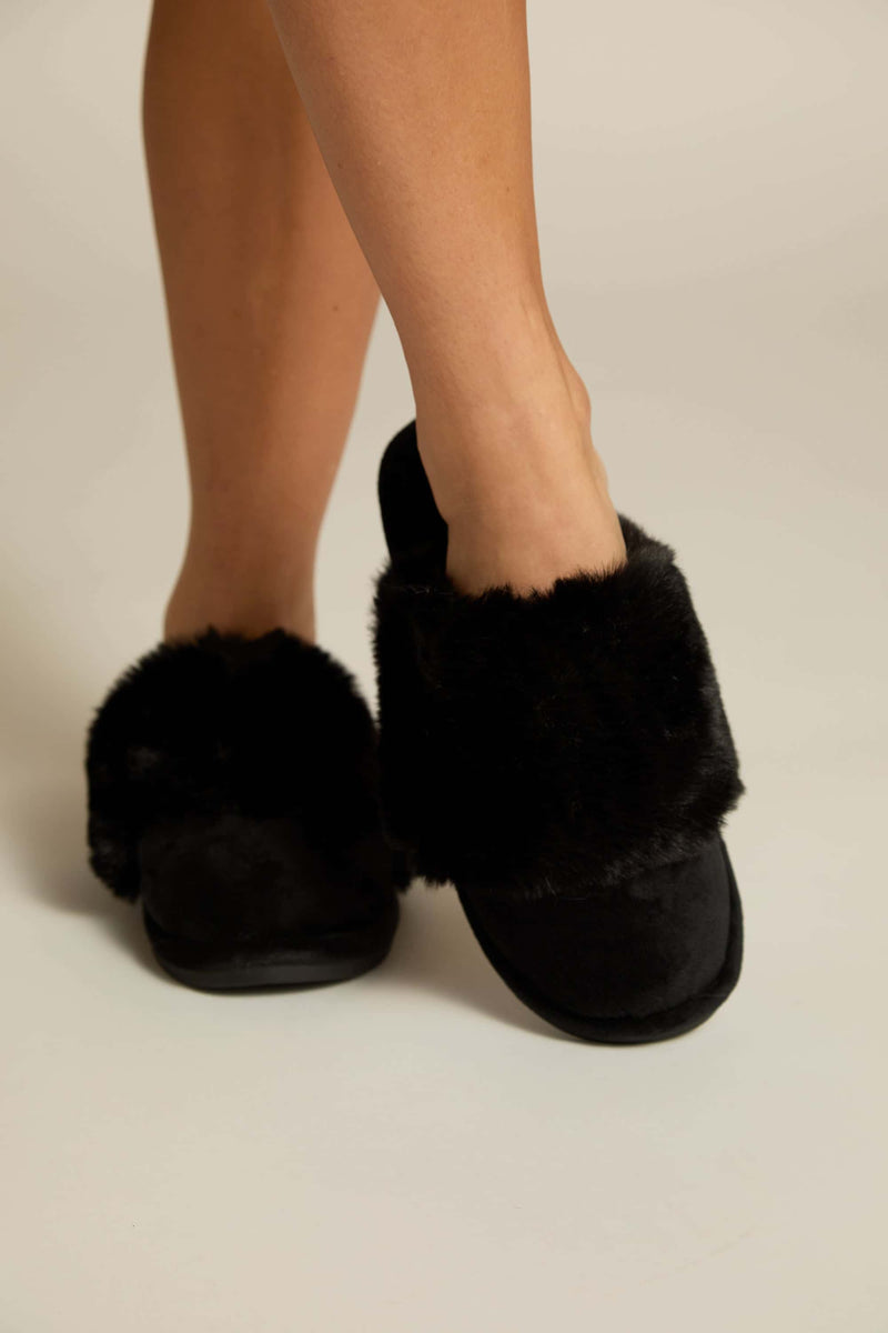 A pair of black furry slippers worn on human feet, showing the plush upper and the sole 