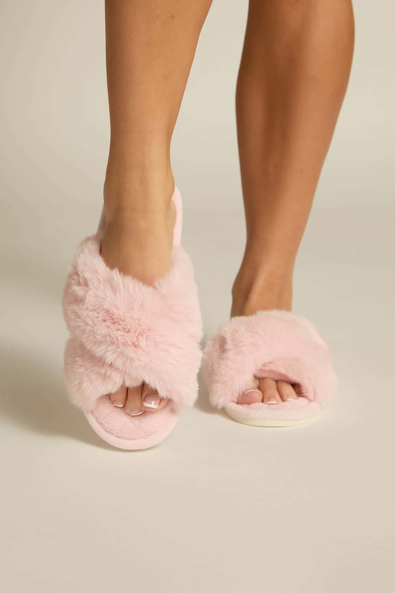A pair of pink furry slides on a person's feet, designed like house slippers with a plush upper and open-toe design.