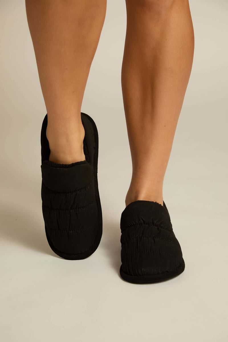 A pair of black puffy slippers worn on a person's feet against a light background.