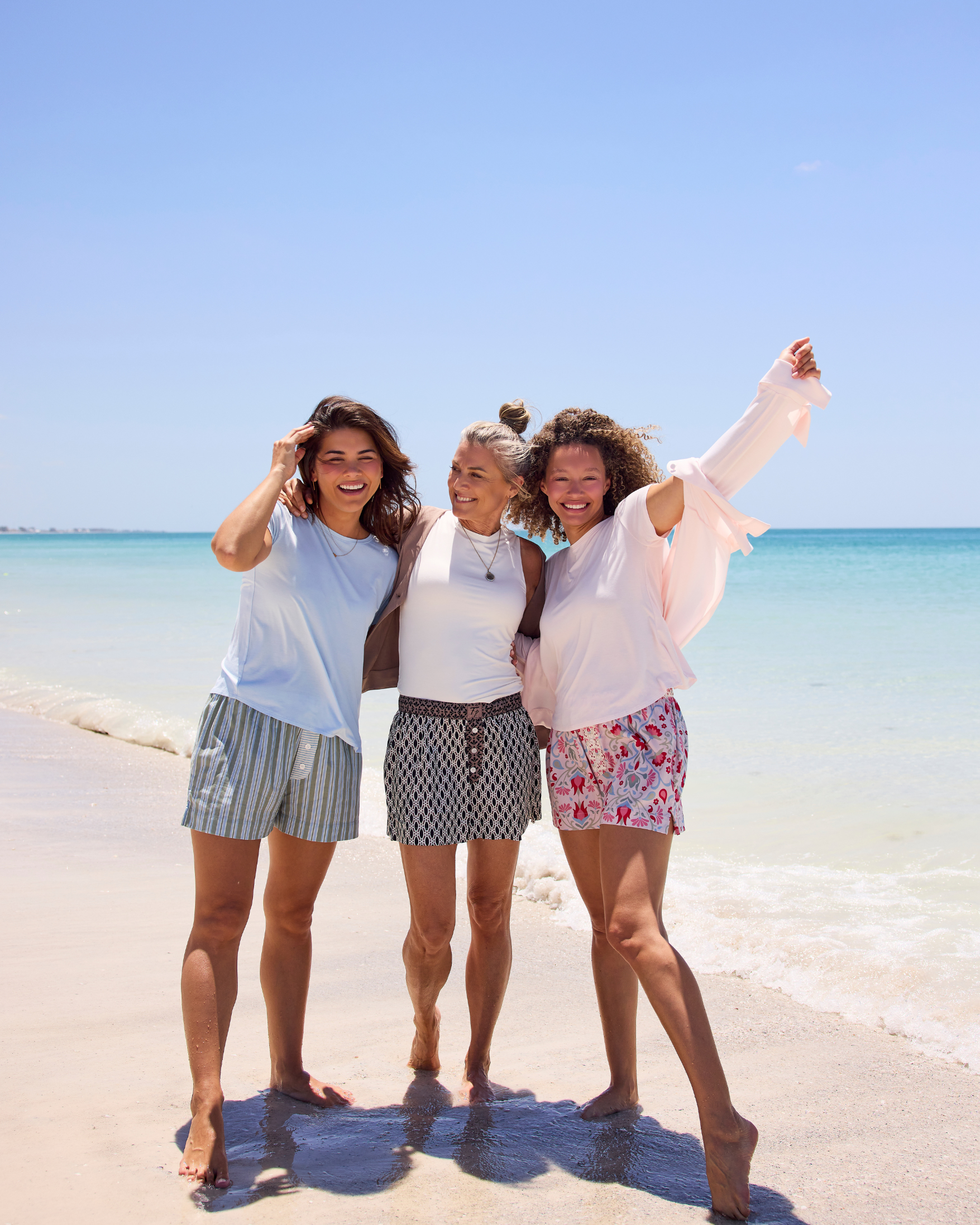 Three women on a sunny beach with the ocean and blue sky in the background, wearing Faceplant Dreams' new cotton print shorts and the softest breathable loungewear tops in white, sky blue, and pink.