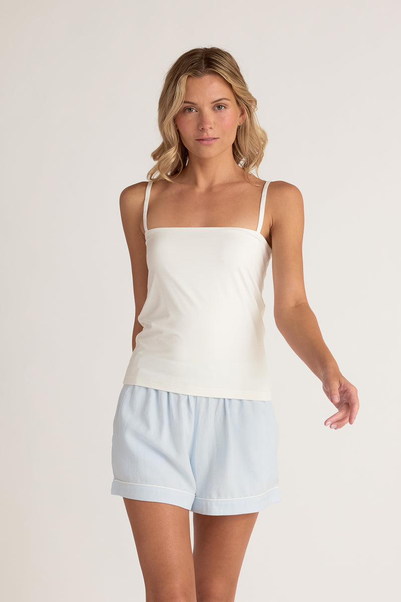 Woman wearing a white tank top and light blue pajama shorts on a plain background