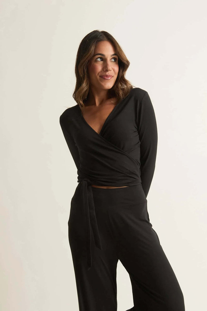 A woman wearing a black Studio Wrap Top with long sleeves, featuring a crossover neckline and tie-to-fit in front or back. The top is made of bamboo and has a cozy, breathable texture.