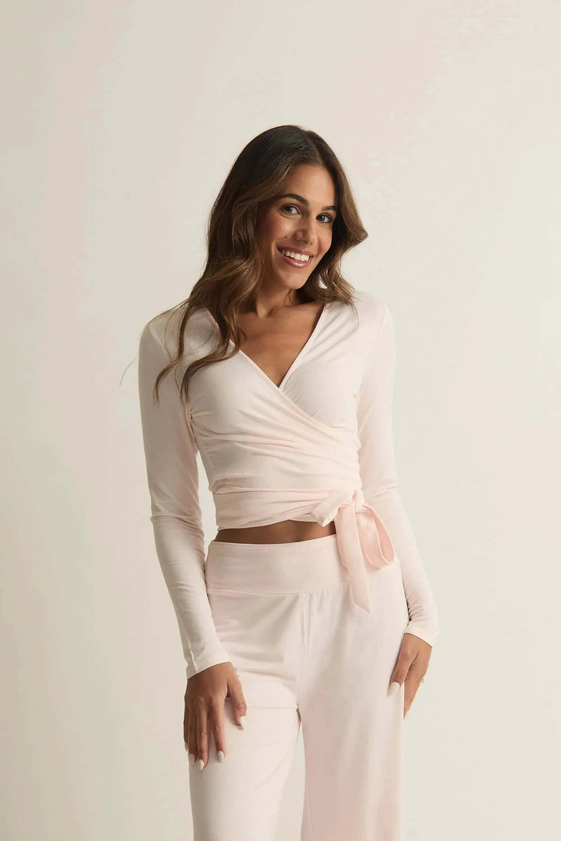 A woman wearing a pink Studio Wrap Top with long sleeves, featuring a crossover neckline and tie-to-fit in front or back. The top is made of bamboo and has a cozy, breathable texture.